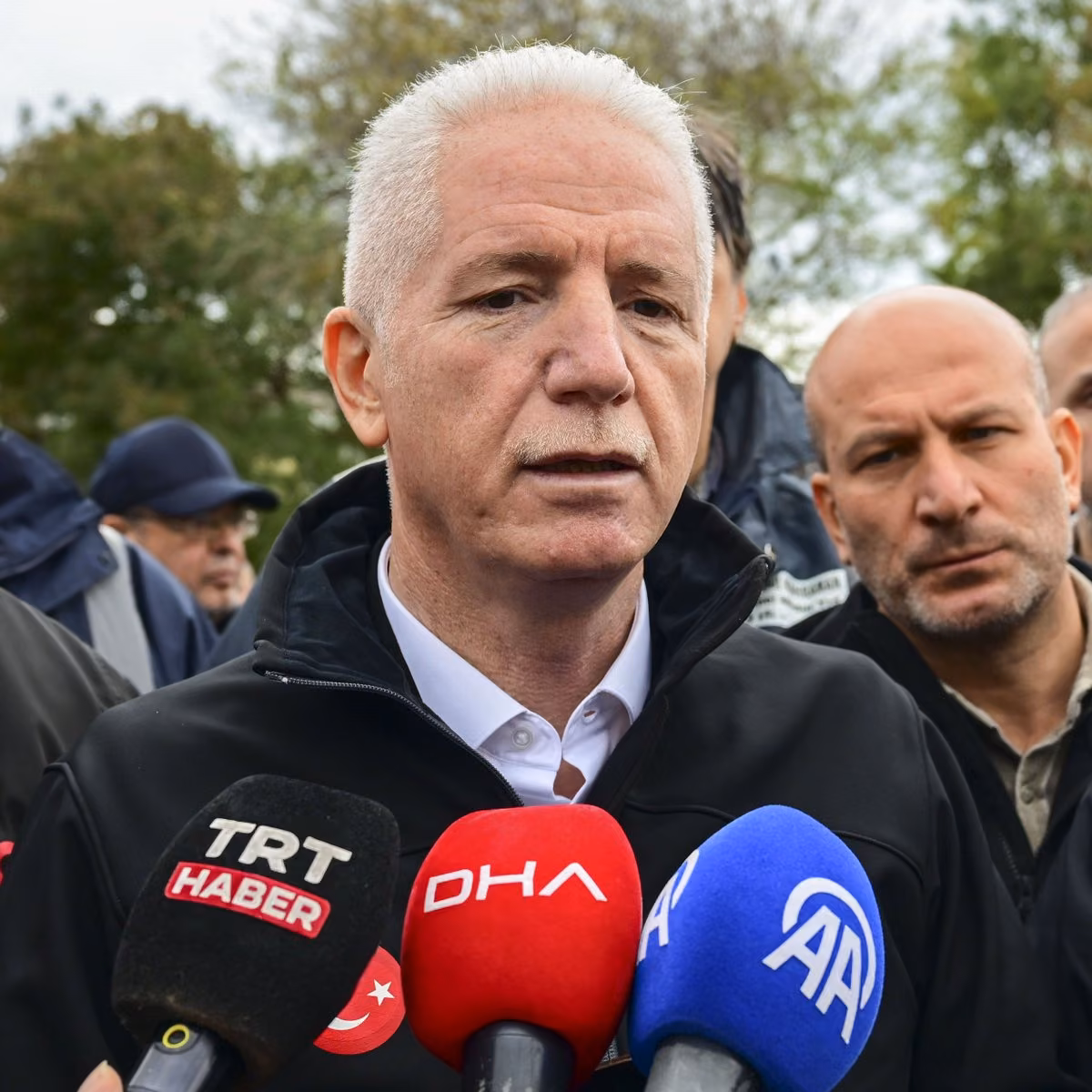 Beyaz saçlı, siyah mont giyen bir erkek; önünde TRT Haber, DHA ve AA mikrofonları var, açıklama yapıyor.
