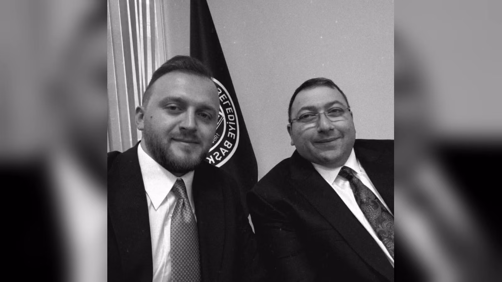 Takım elbiseli iki adam, arkalarında amblemli koyu renk bir bayrakla siyah beyaz bir karede yan yana poz veriyor.