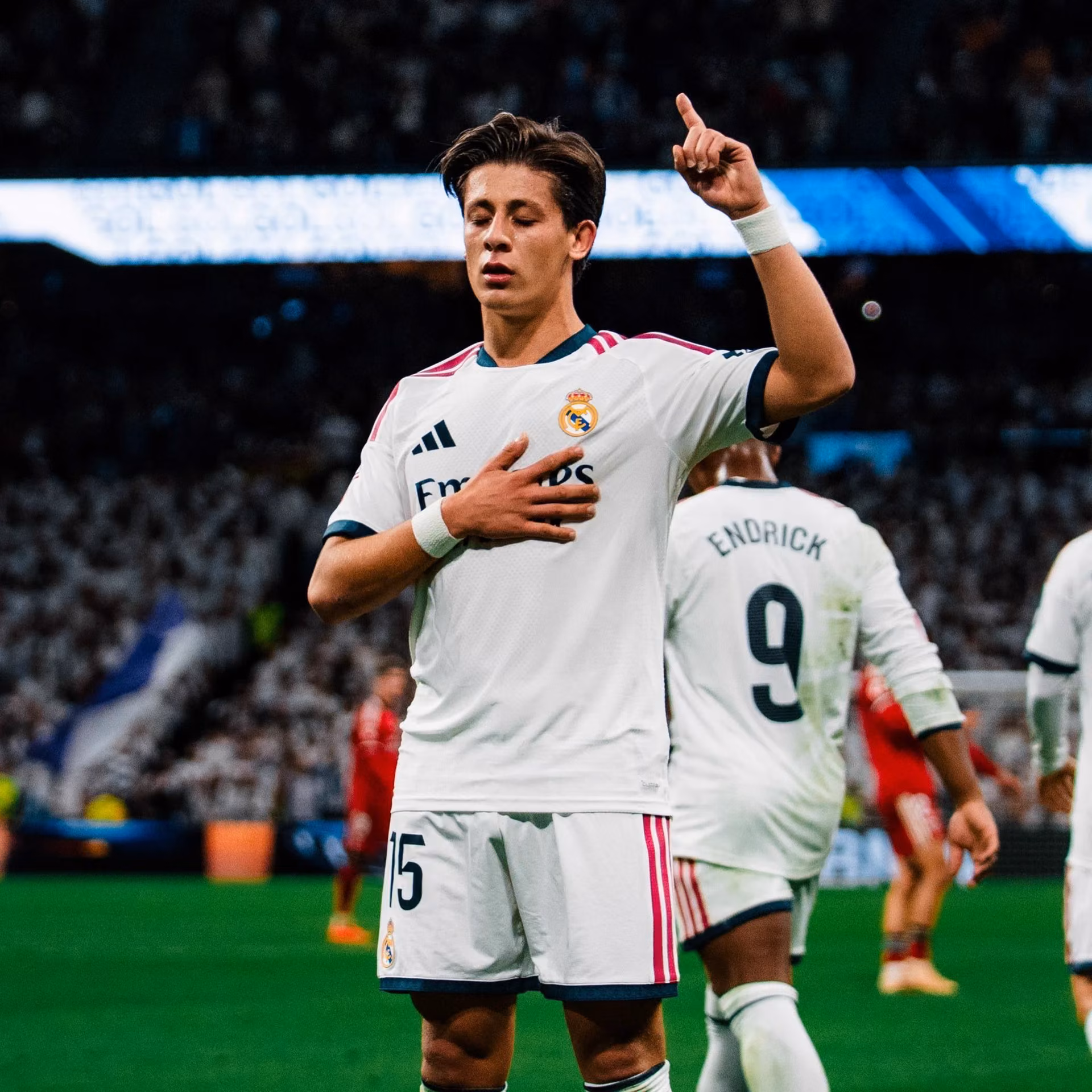 Beyaz Real Madrid forması giyen, elini göğsüne koyup parmağını havaya kaldıran bir futbolcu stadyumda kutlama yapıyor.