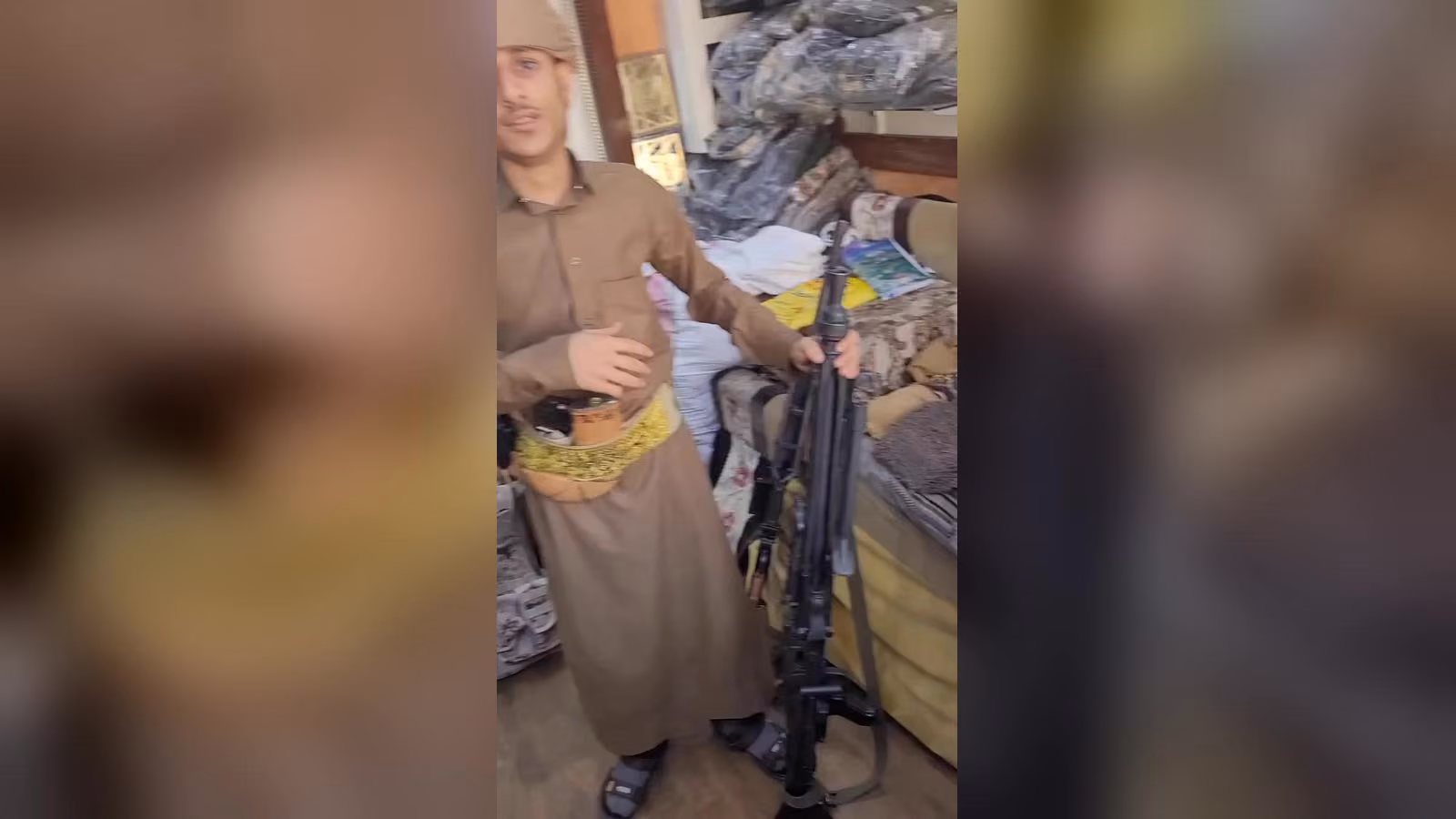 Kahverengi geleneksel kıyafetli bir adam, yatak ve battaniyelerin olduğu odada makineli tüfek tutuyor.