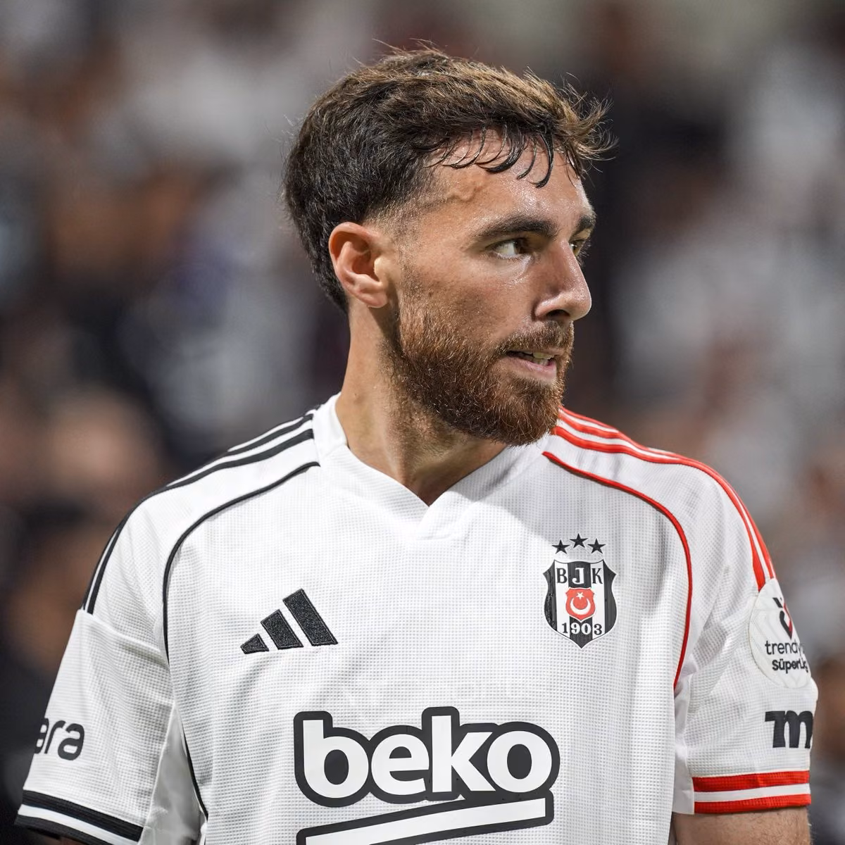 Beyaz-siyah-kırmızı Beşiktaş forması giyen, sakallı ve kısa kahverengi saçlı bir futbolcu yan tarafa bakıyor. Arka planda bul