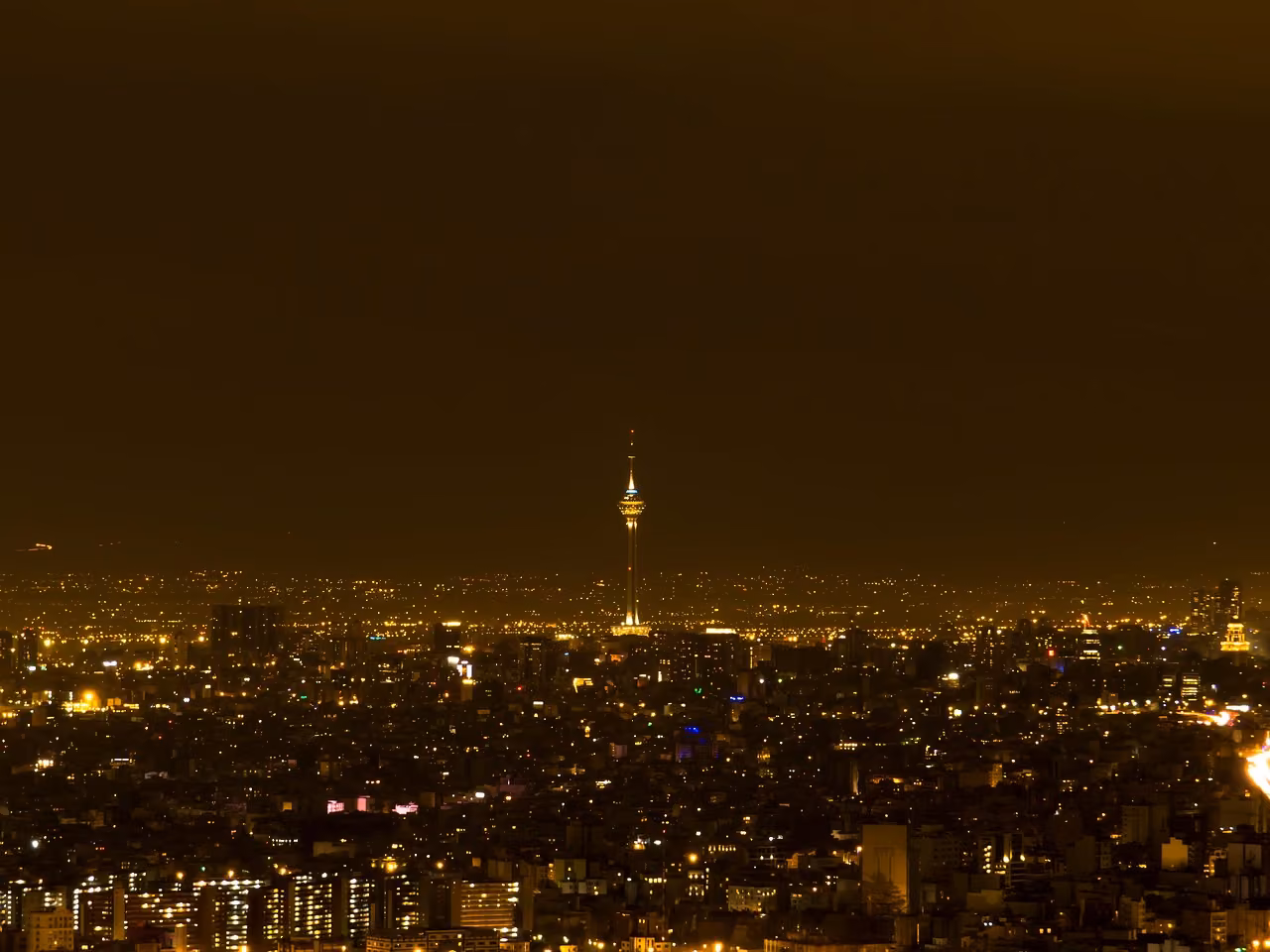 Gece karanlığında yoğun ışıklarla aydınlanmış geniş bir şehir manzarası; ortada ince uzun bir kule yükseliyor.