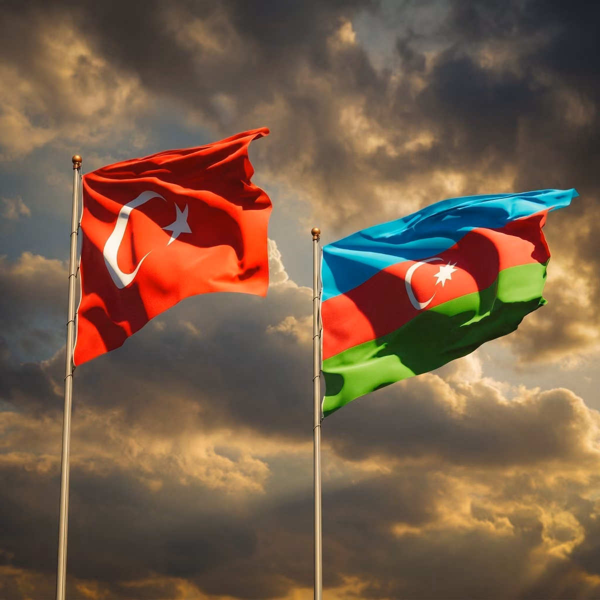Dramatik bulutlu gökyüzü altında yan yana dalgalanan kırmızı Türk bayrağı ve mavi-kırmızı-yeşil Azerbaycan bayrağı.