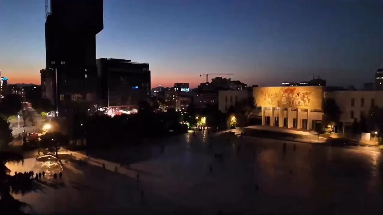 Akşam karanlığında geniş bir meydan; sağda kabartmalı, aydınlatılmış bir müze binası, solda yüksek karanlık bir bina ve birka