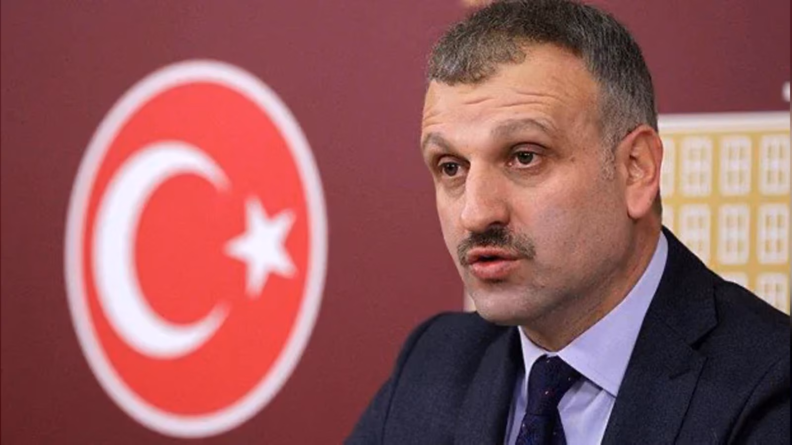 Koyu takım elbiseli, kravatlı, bıyıklı bir erkek; arkasında kırmızı zemin üzerinde ay yıldızlı bir amblem var.
