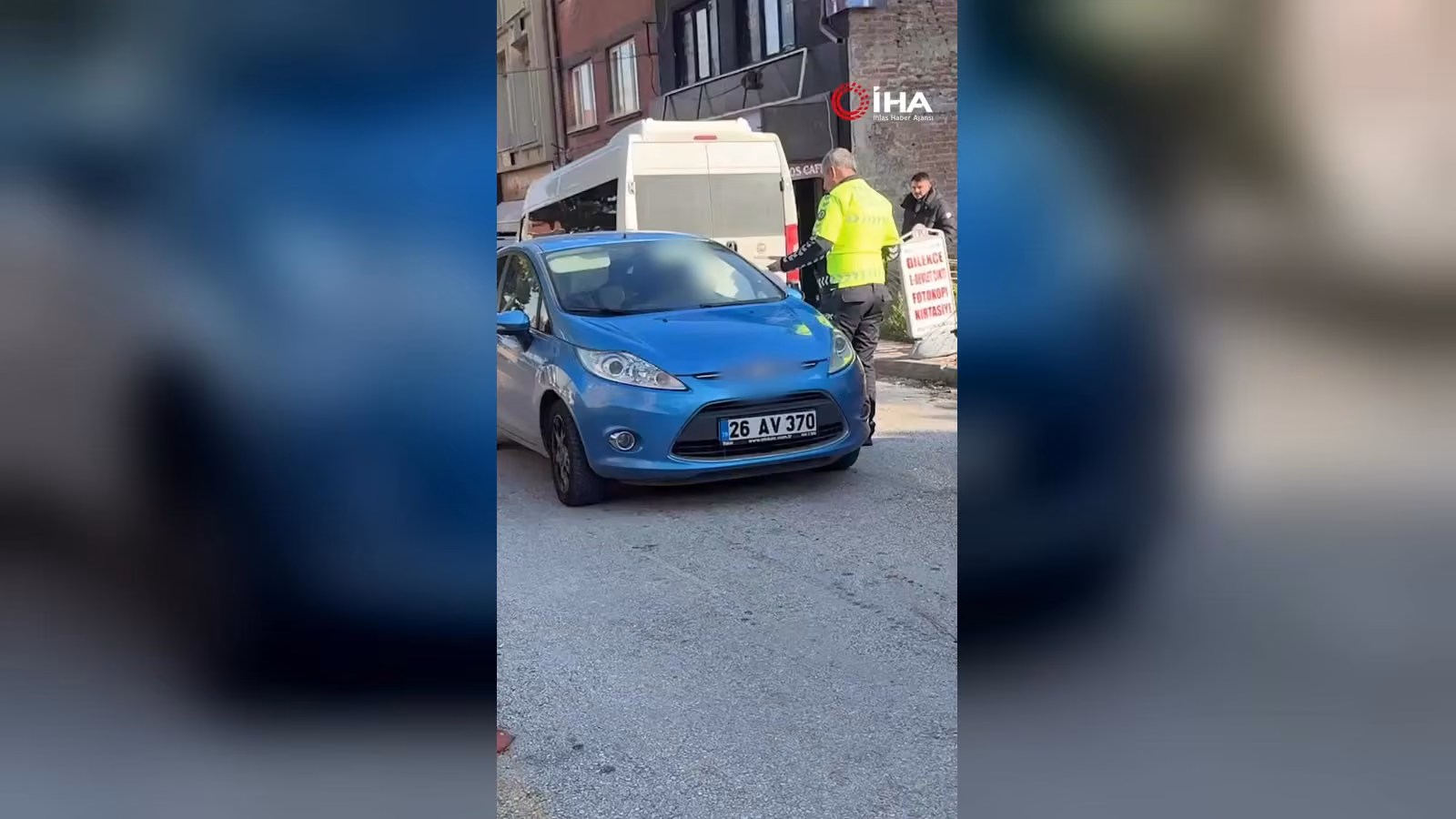 Trafik polisi yeşil yelekle mavi bir otomobilin yanında duruyor; arkada beyaz minibüs ve dükkan tabelaları görünüyor.