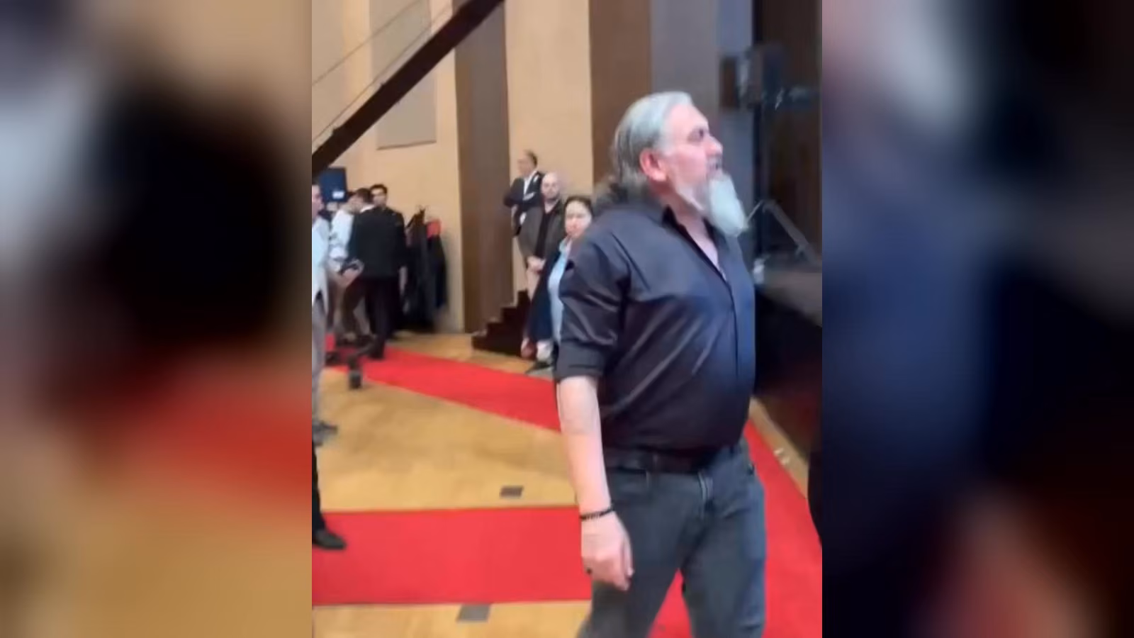 Kırmızı halılı salonda, gri saçlı ve sakallı, koyu gömlekli bir adam sağa doğru yürüyor; arkada birkaç kişi duruyor.