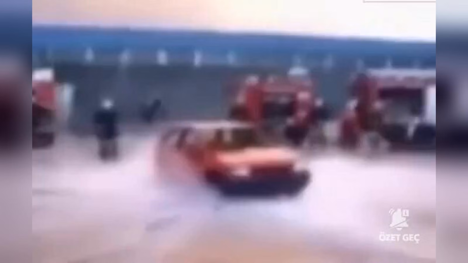 Kırmızı bir otomobil su ve köpük bulutu içinde; arkada kırmızı itfaiye araçları ve birkaç kişi görünüyor.