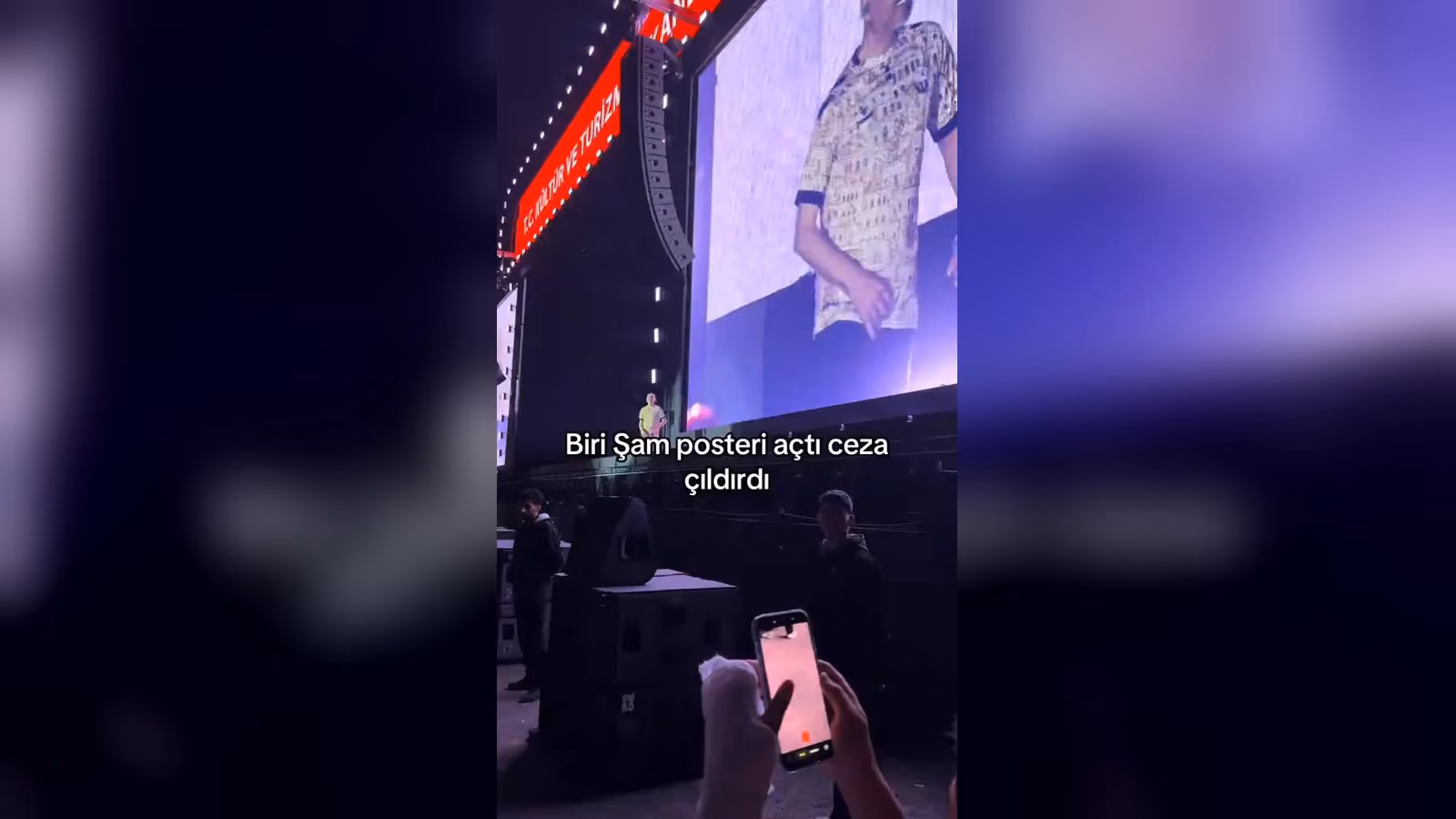 Konser sahnesi; arkada dev ekranda desenli gömlekli bir adam var, önde izleyiciler telefonla kayıt yapıyor.