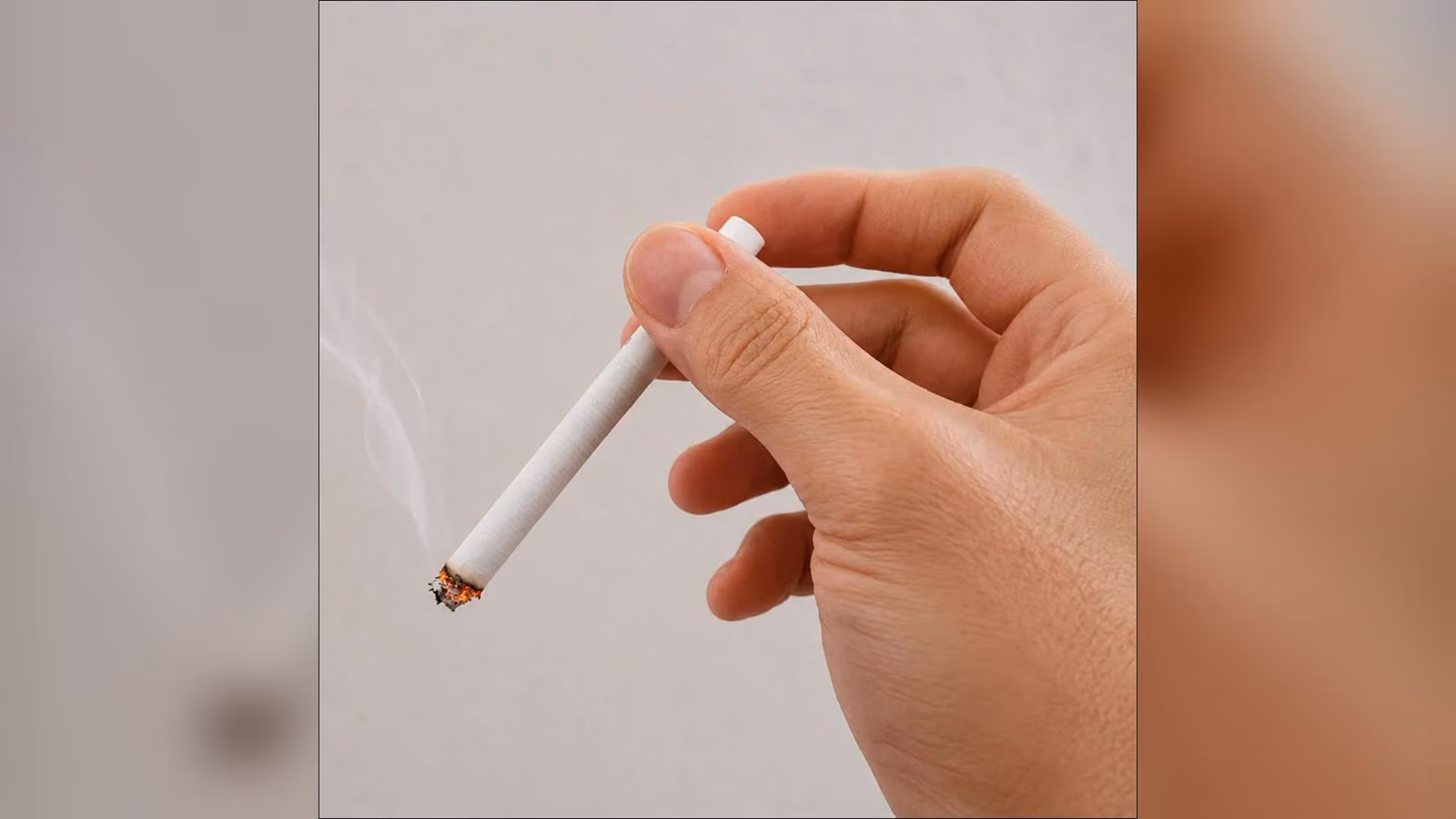 A hand holding a lit cigarette between its fingers, with a thin trail of smoke rising against a plain grey background.