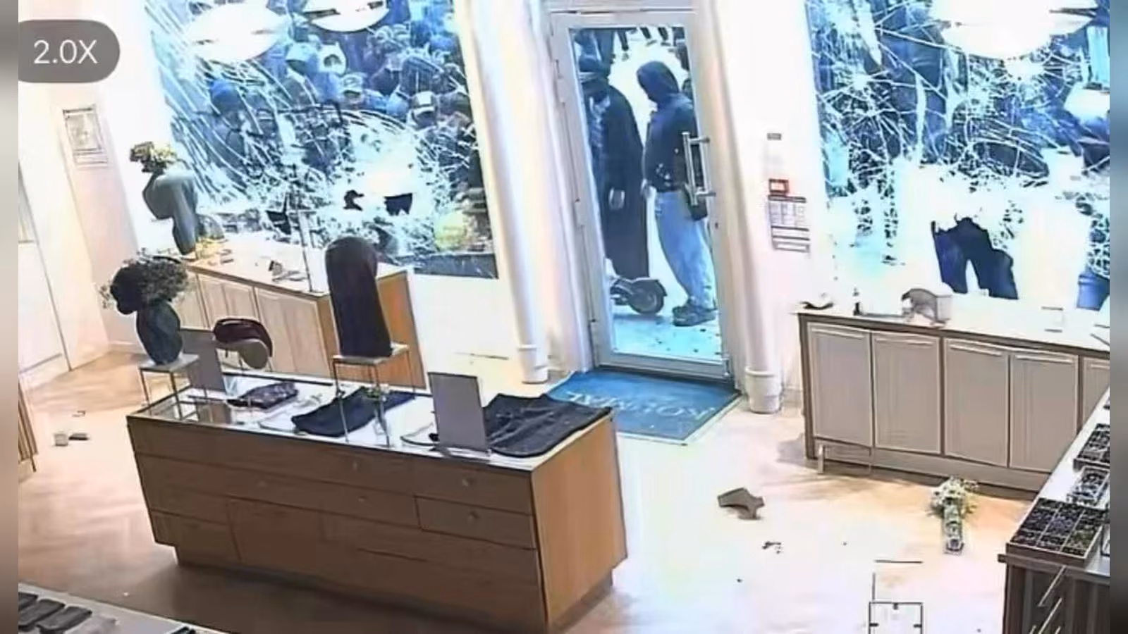 Vitrinleri kırılmış bir mağazanın içi; kapıda kapüşonlu iki kişi duruyor, yerde cam kırıkları ve dağınık eşyalar var.