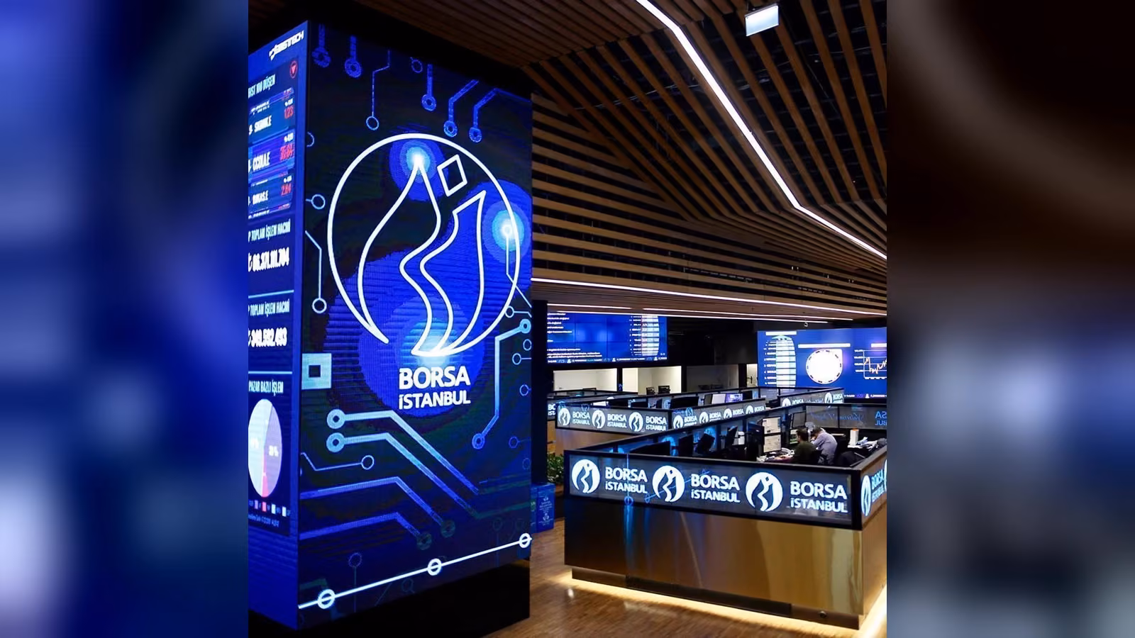 Mavi LED ekranda Borsa İstanbul logosu; arkada bilgisayar masalarında çalışan kişilerin olduğu bir işlem salonu.