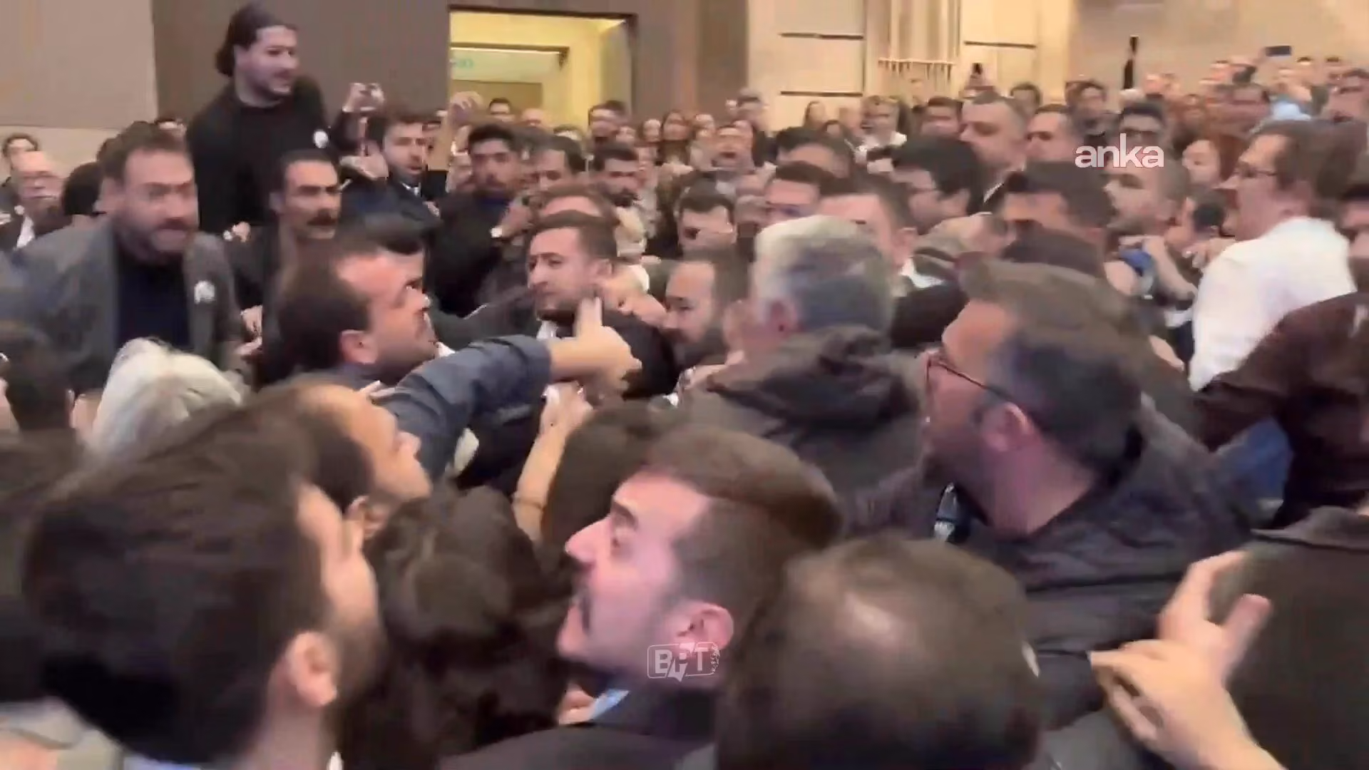 Kalabalık bir salonda birçok kişi sıkışmış durumda; ortadaki bir adam parmağıyla karşısındakine işaret ediyor, çevredekiler i