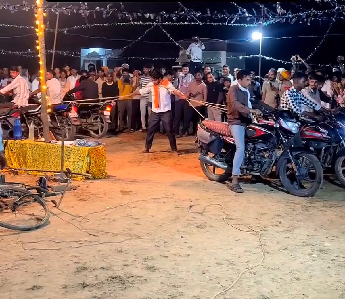 Gece meydanda, turuncu atkılı bir adam iplerle sıradaki motosikletleri çekiyor; kalabalık izliyor.