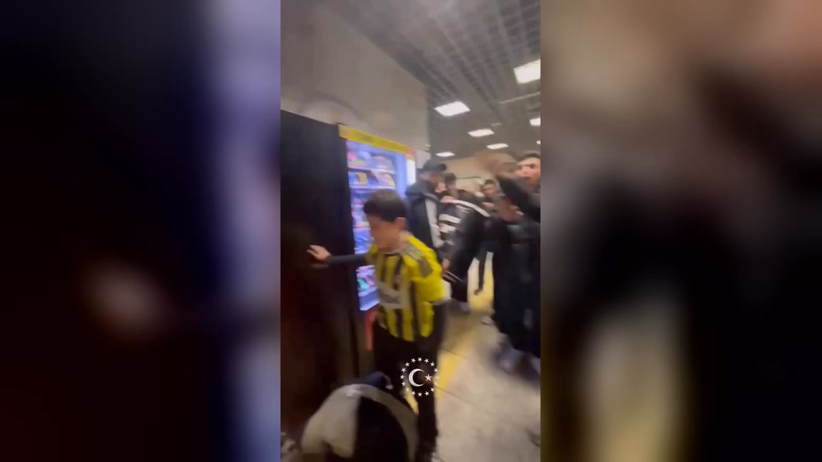 Metroda sarı-siyah çizgili Fenerbahçe formalı bir çocuk, yanında kalabalık bir grup; arka planda otomat ve lambalar.