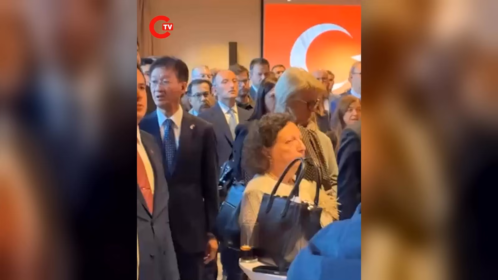 Kalabalık resepsiyonda takım elbiseli, gözlüklü bir erkek ve davetliler; arkada kırmızı zeminde beyaz hilal.