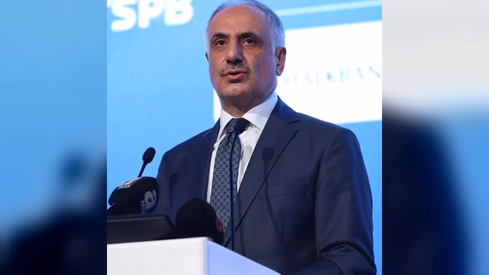 Koyu lacivert takım elbiseli, desenli kravat takmış bir erkek, mavi arka planlı bir kürsünün başında konuşuyor.