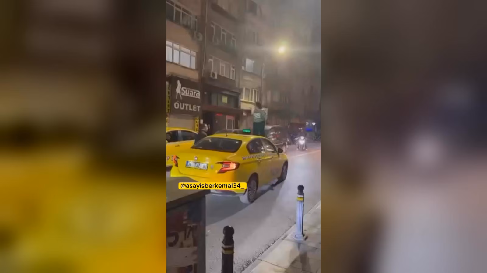 Gece bir caddede sarı bir taksinin tavanında duran kişi; çevrede park halinde araçlar ve binalar var.