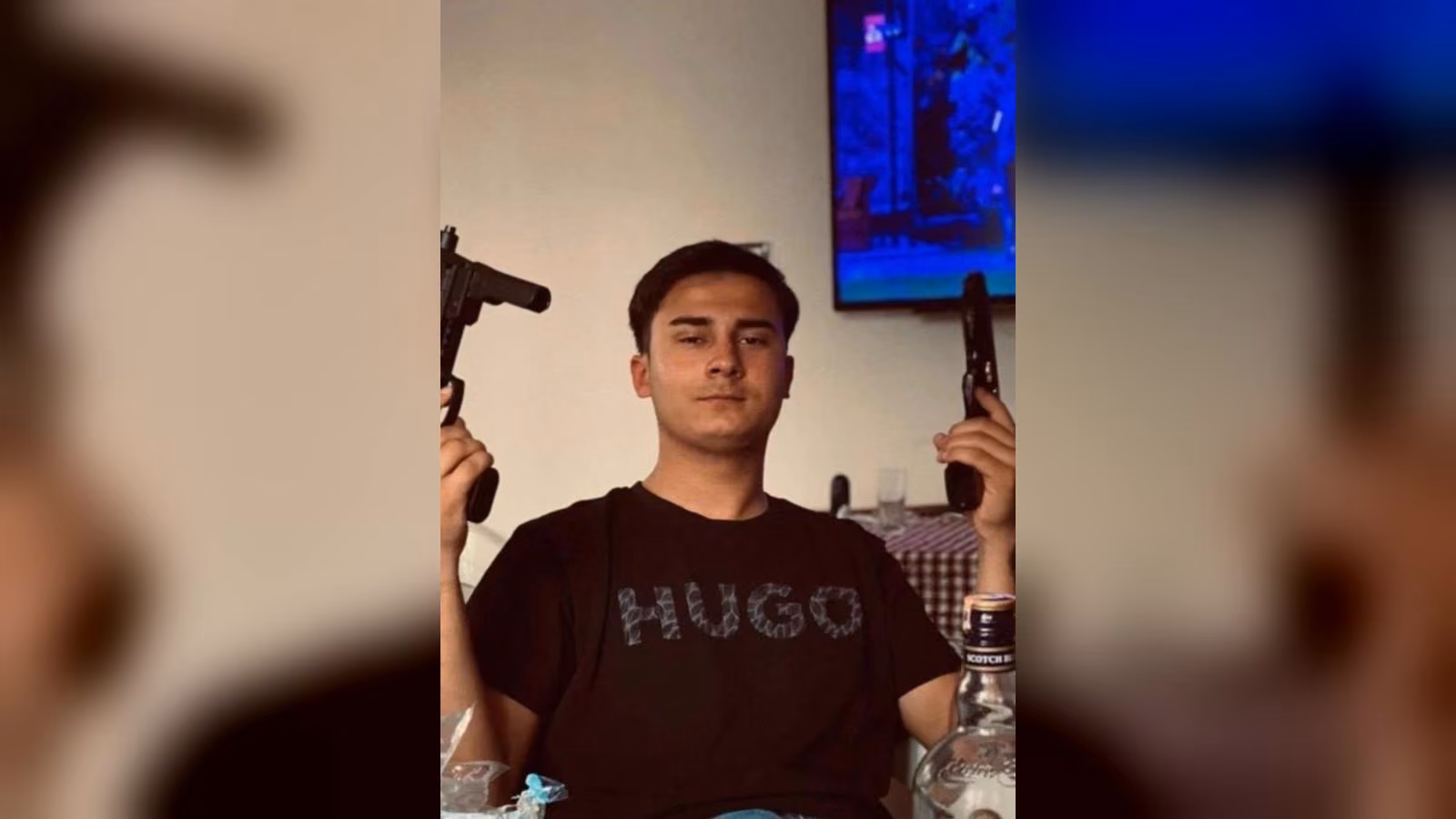 Siyah HUGO tişörtlü genç bir erkek iki elinde tabanca tutuyor; önünde viski şişesi, arkada açık bir televizyon var.