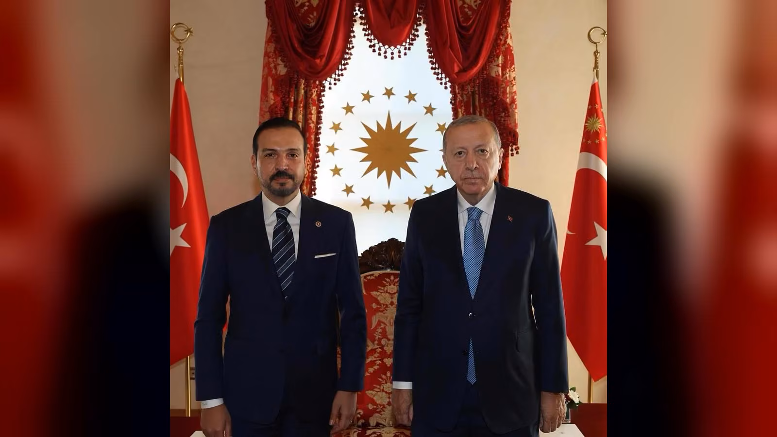 Türk bayraklarının ve kırmızı perdelerin önünde, koyu lacivert takım elbiseli iki erkek yan yana ayakta duruyor.