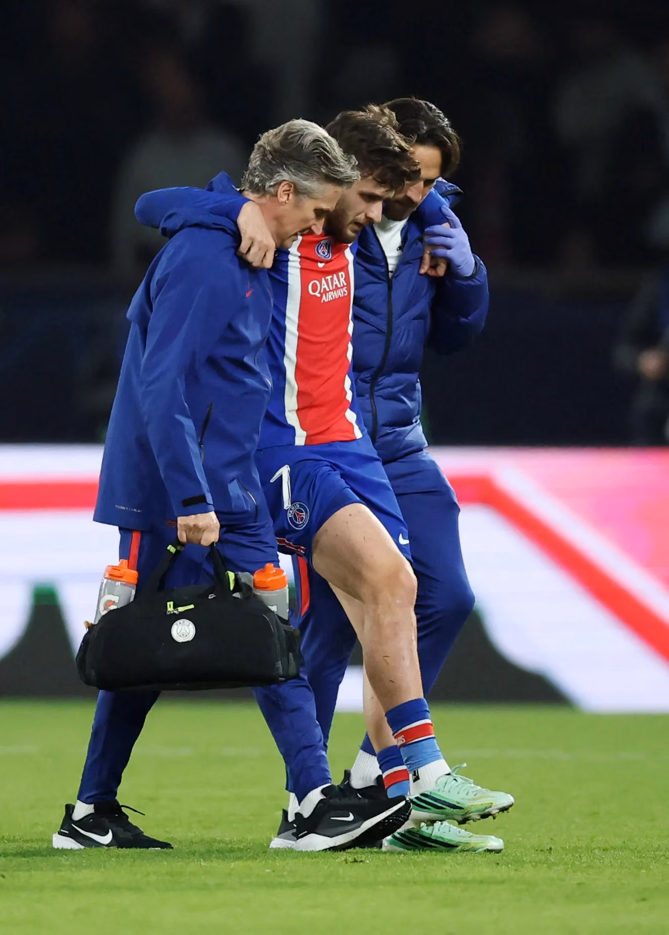 Kırmızı, beyaz ve mavi PSG formalı bir oyuncu, iki sağlık görevlisinin desteğiyle gece sahadan ayrılıyor.
