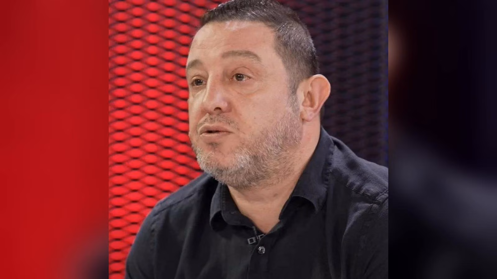Kırmızı dokulu arka planda konuşan; kısa saçlı, sakallı, siyah gömlekli bir erkek. Yakasında yaka mikrofonu var.