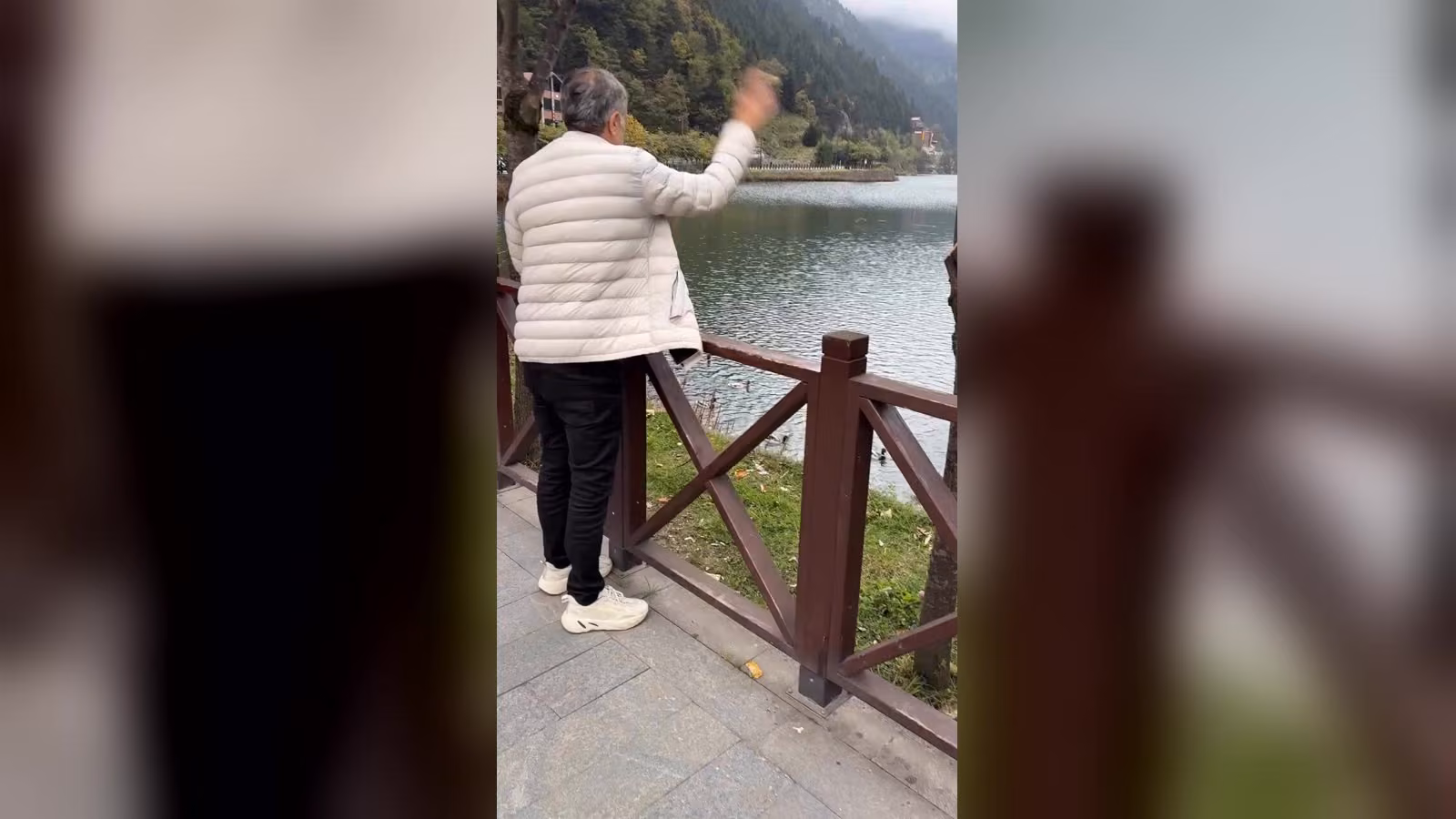 Beyaz şişme mont giyen bir adam, Uzungöl'de ahşap korkuluğun yanında elini sallıyor. Önünde göl ve ormanlık dağlar var.