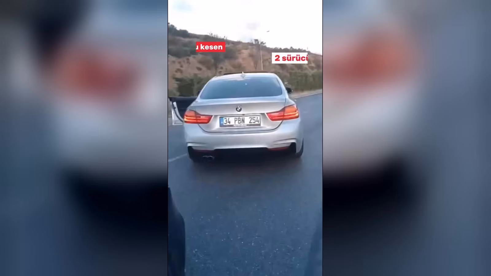 Dağlık bir yolda ilerleyen gümüş rengi BMW sedanın arkadan görünümü; görüntüde kırmızı etiketli yazılar var.