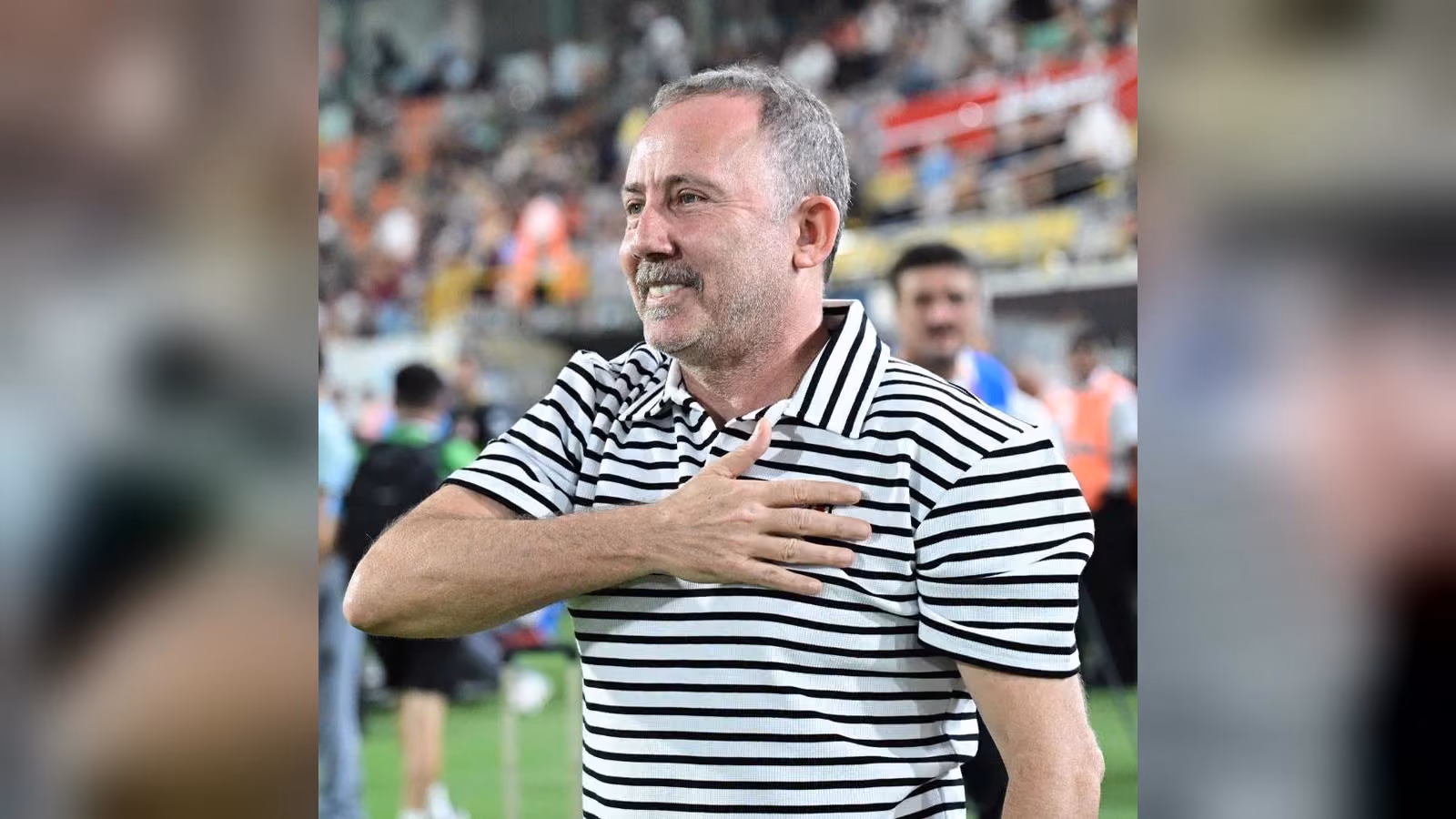 Çizgili beyaz-siyah polo gömlekli, gri saçlı bir erkek stadyumda gülümseyerek elini göğsüne koymuş.