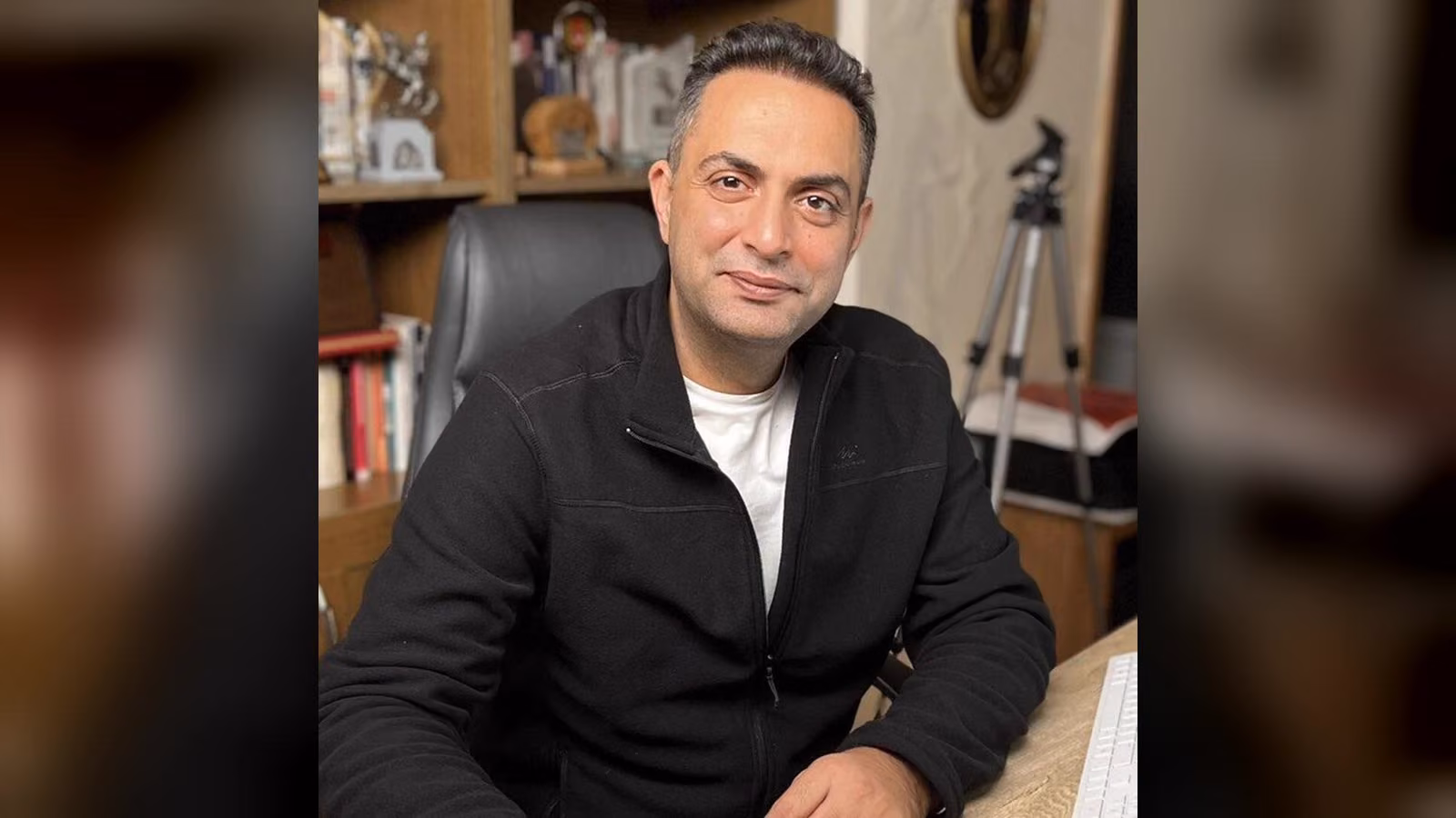 Siyah fermuarlı ceket giyen gri saçlı bir adam, kitaplıklı bir odada masa başında oturmuş hafifçe gülümsüyor.