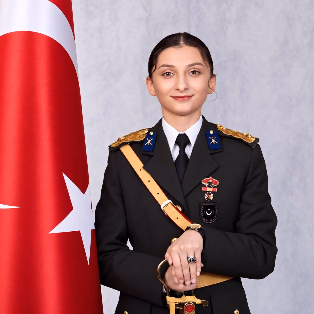 Türk bayrağının yanında, altın rengi omuz ve kuşak aksesuarlı koyu askeri üniformalı, gülümseyen genç bir kadın.