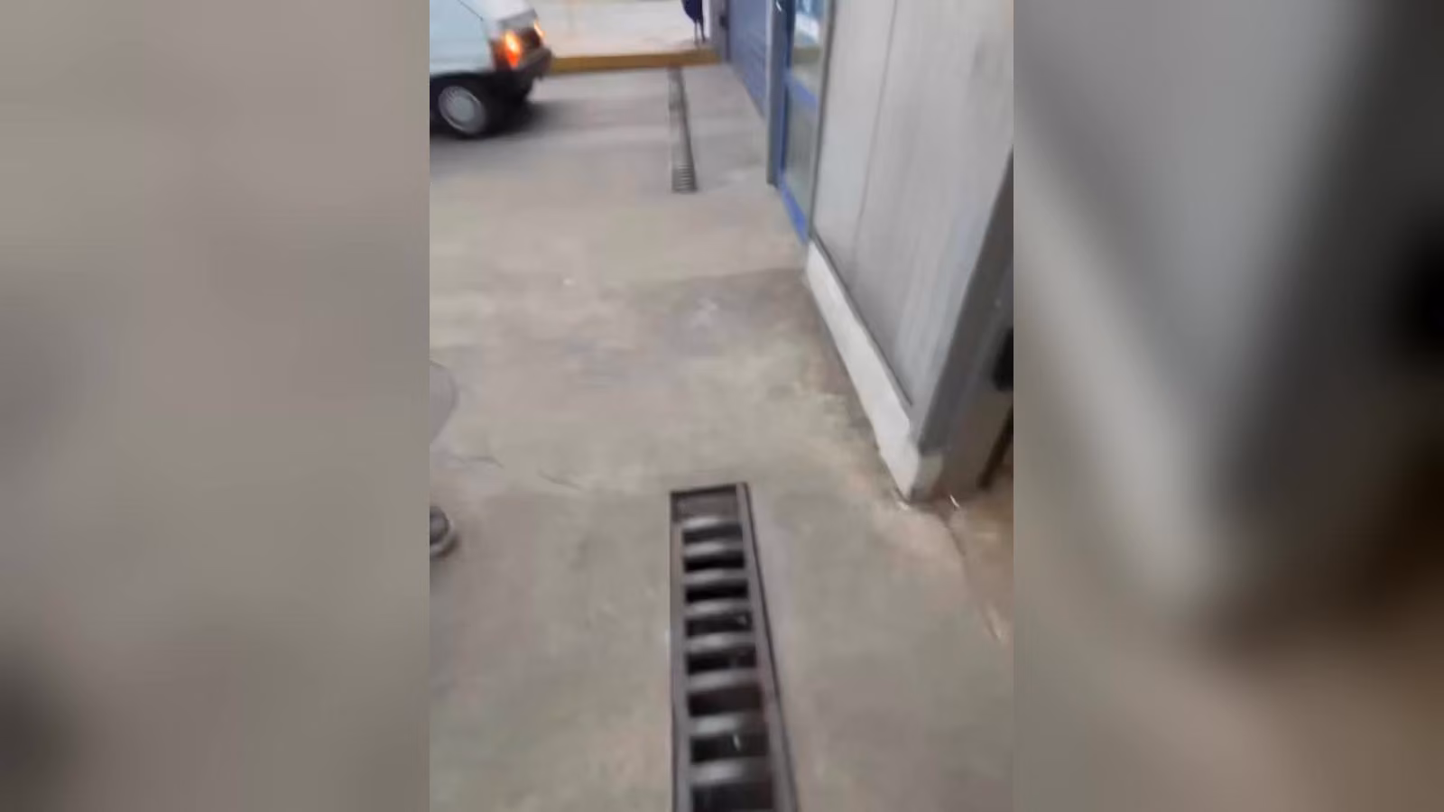 Beton zeminde metal ızgaralı kanal; solda stop lambası yanan beyaz bir van ve mavi çerçeveli camlı bir binanın girişi.