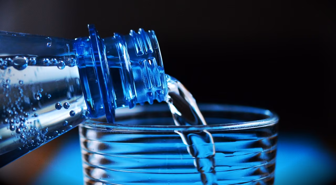 Mavi ışıkla aydınlatılmış plastik su şişesinden cam bardağa su dökülüyor; sudaki kabarcıklar ve koyu arka plan görünüyor.