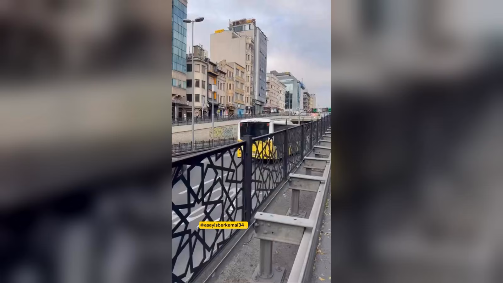 Siyah süslü korkuluğun arkasında, yolda ilerleyen sarı-beyaz bir otobüs ve arkasında çok katlı binalar