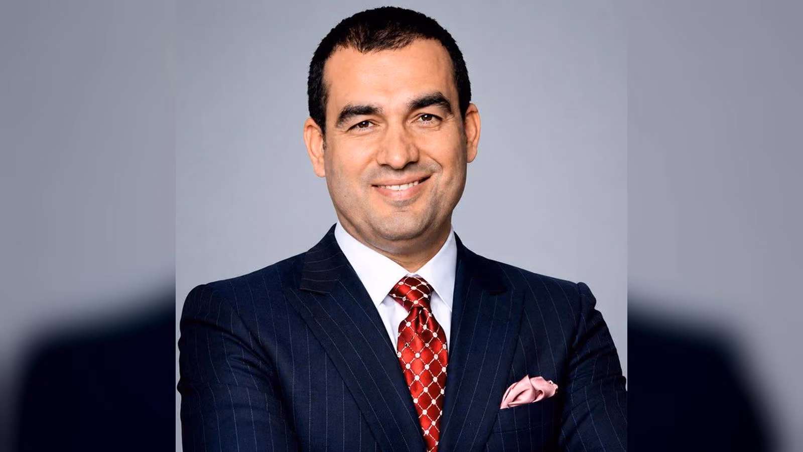 Lacivert çizgili takım elbiseli, kırmızı desenli kravatlı, gülümseyen bir erkek; gri fonda kollarını bağlamış.