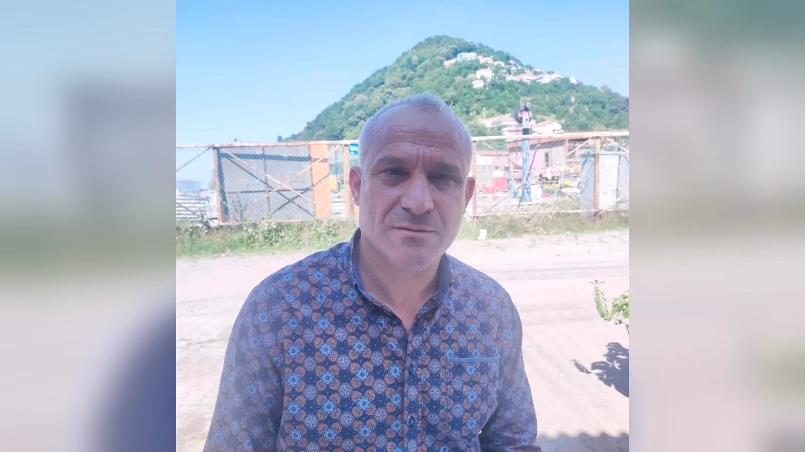 Desenli mavi ve kahverengi gömlekli, kısa saçlı bir adam açık havada duruyor; arkasında yeşil ağaçlı bir tepe ve yapılar var.