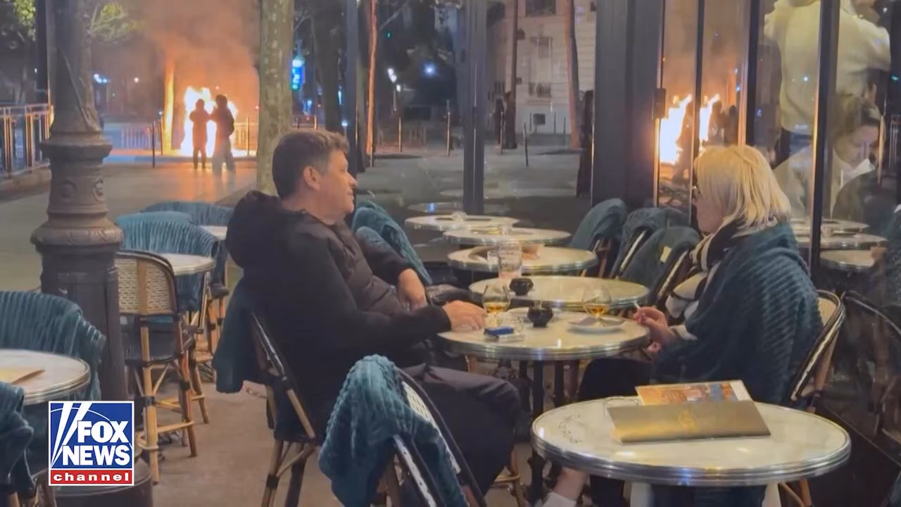 Gece vakti bir kafe terasında oturan koyu renk ceketli bir erkek ve sarışın bir kadın; arkada sokakta yanan bir çöp kutusu