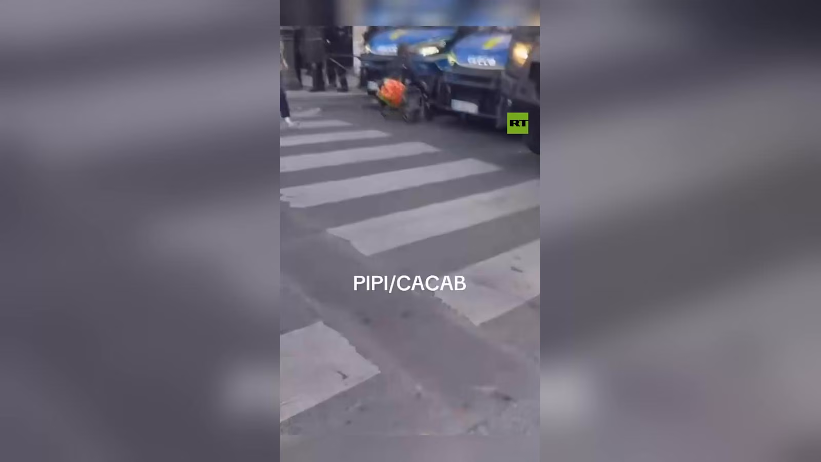 Şeritli yaya geçidi ve arkasında park etmiş mavi polis araçları; önde turuncu poşetli tekerlekli bir sepet görülüyor.