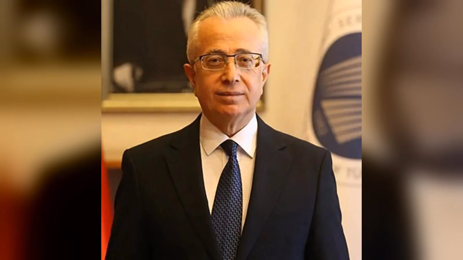 Gözlüklü, gri saçlı bir erkek koyu lacivert takım, beyaz gömlek ve desenli kravatla bayrak önünde duruyor.