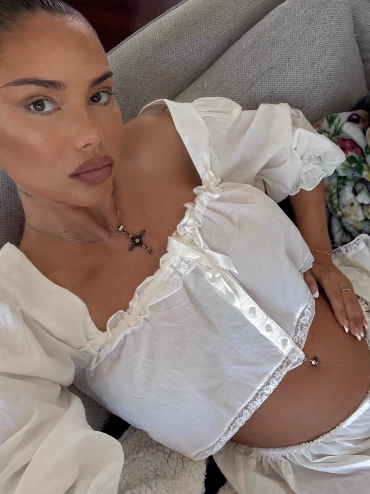 Beyaz dantelli bluzlu, kanepede uzanmış halde selfie çeken bir kadın; yıldız figürlü kolye takıyor.