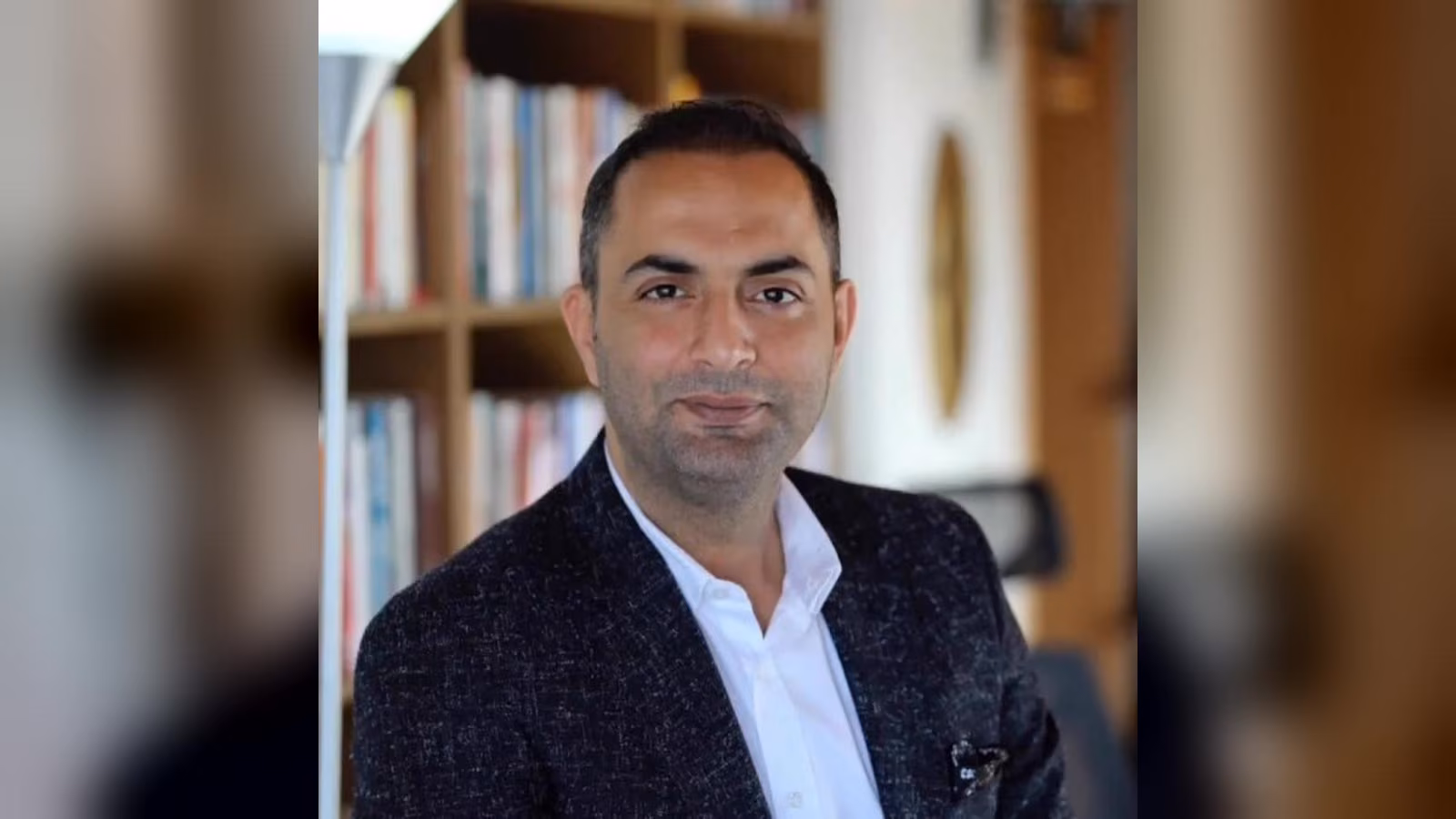 Koyu renk desenli ceket ve beyaz açık yakalı gömlek giymiş, kısa saçlı bir erkek; arkasında kitaplıklar ve bir lamba var.
