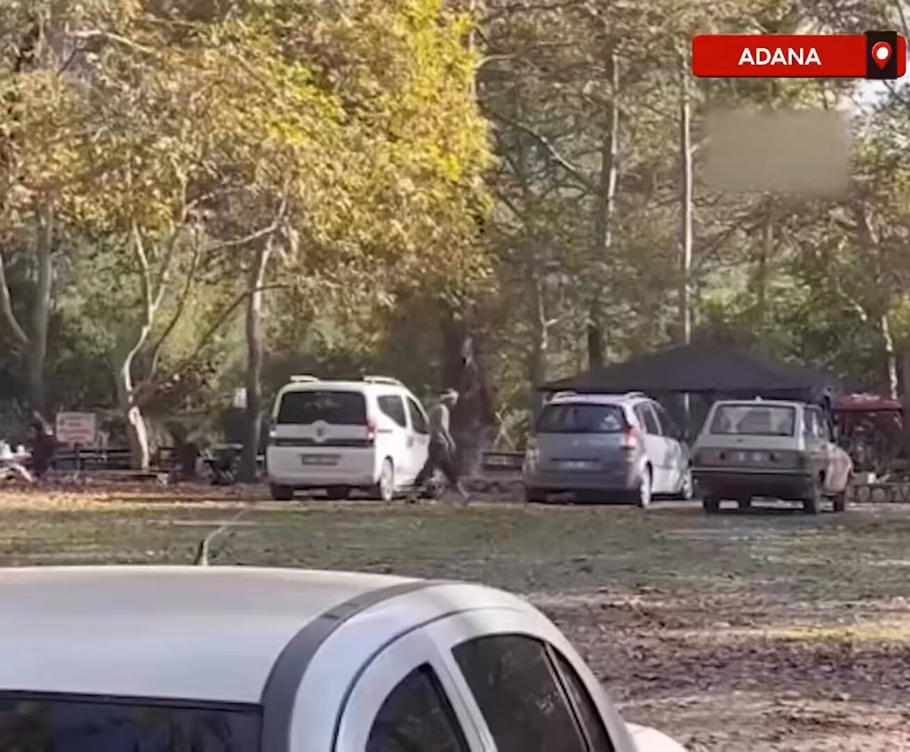 Ağaçlı bir park alanında beyaz minibüs ve park etmiş araçların yanından koyu kıyafetli bir kişi yürüyor.