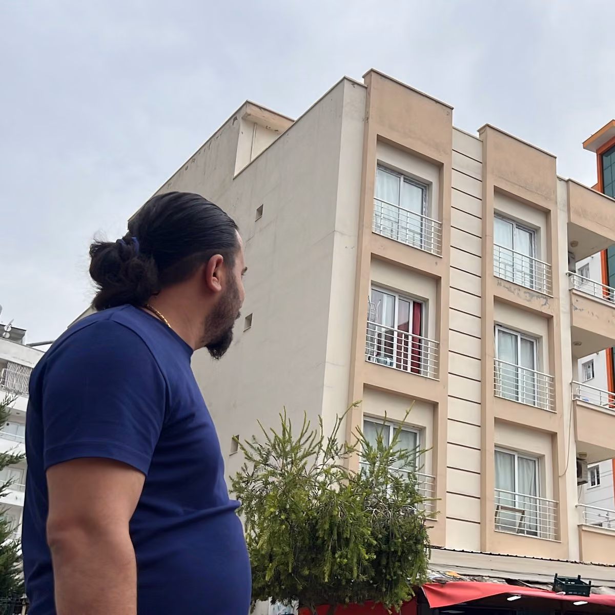 Lacivert tişörtlü, saçı topuz yapılmış bir erkek, bej renkli çok katlı binanın balkonlu cephesine bakıyor.