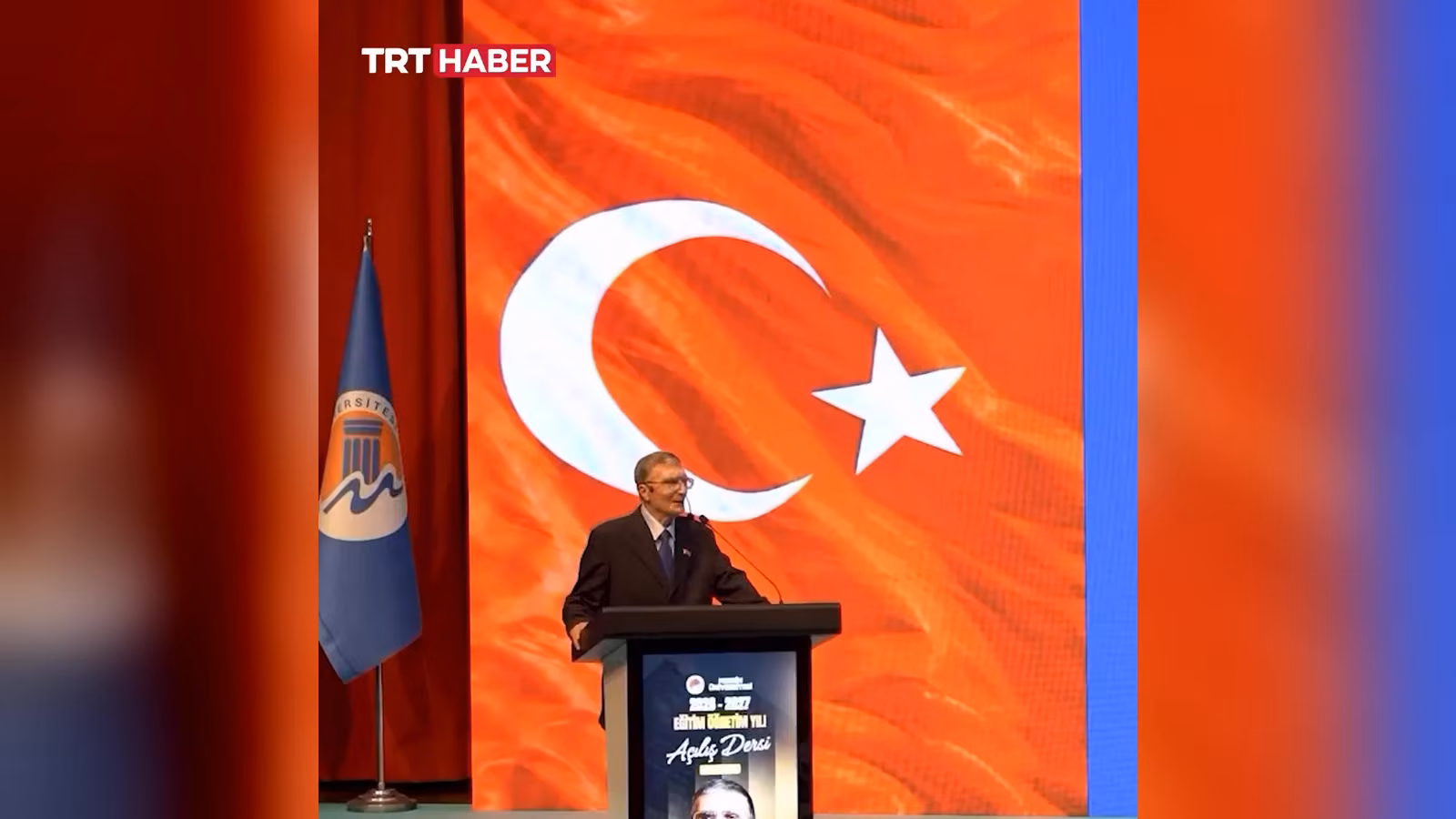 Kürsüde konuşan bir erkek. Arkasında büyük bir Türk bayrağı ve solda Mersin Üniversitesi bayrağı var.