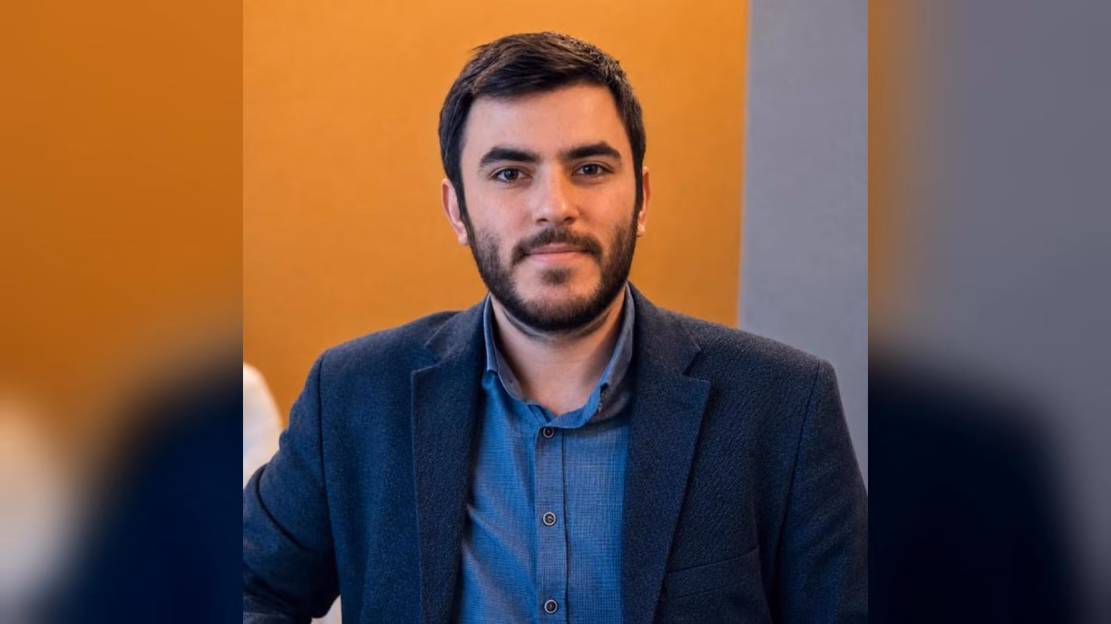 Koyu mavi ceketli, mavi gömlekli, kısa saçlı ve sakallı bir erkek turuncu fonun önünde kameraya bakıyor.