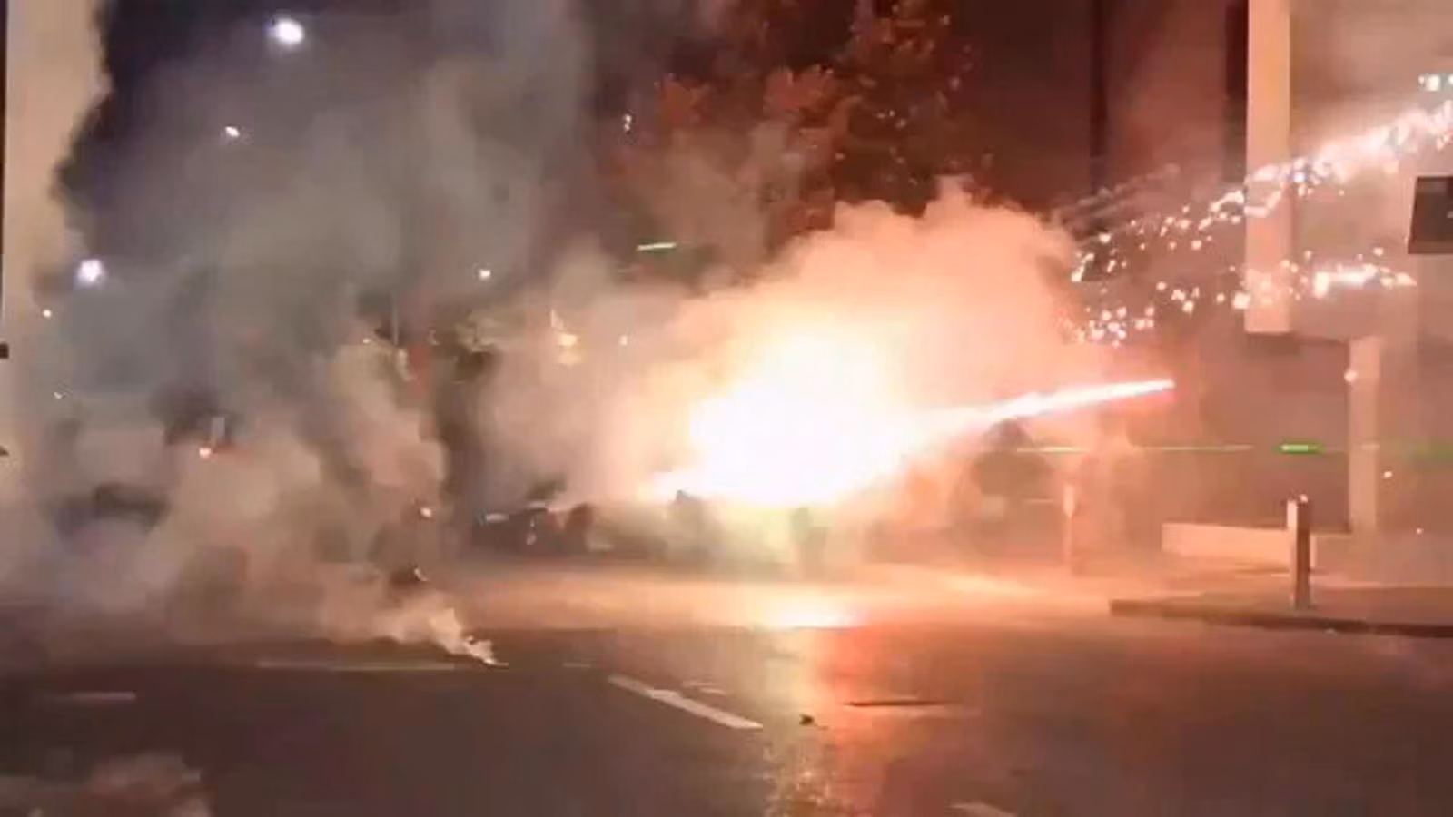 Gece bir caddede, dumanın içinden parlayan havai fişek ve etrafa saçılan kıvılcımlar görünüyor.