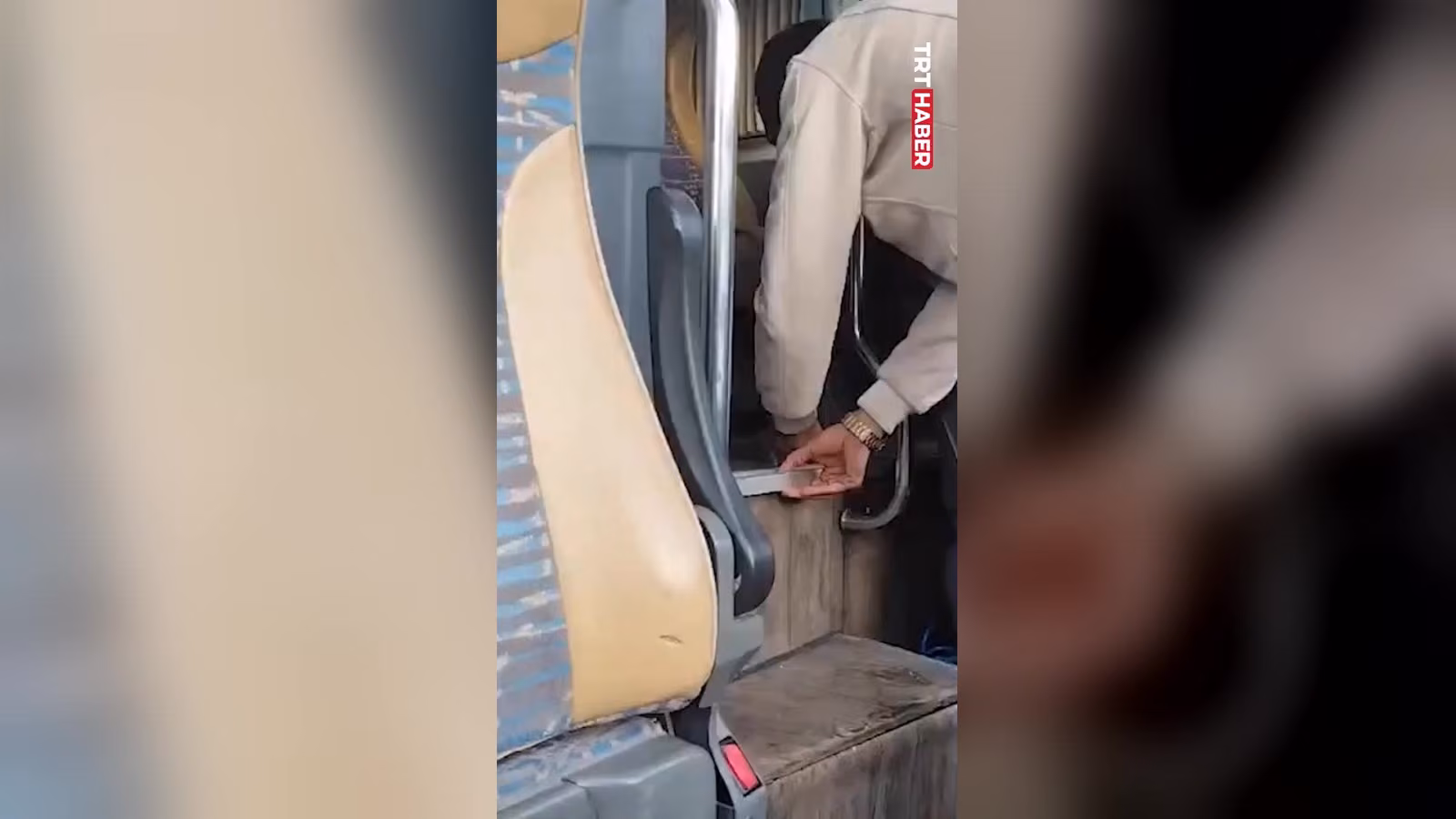 Otobüs koltuğunun yanında, bej ceketli bir adam zemindeki ahşap kapaklı bir bölmeyi açmaya çalışıyor.
