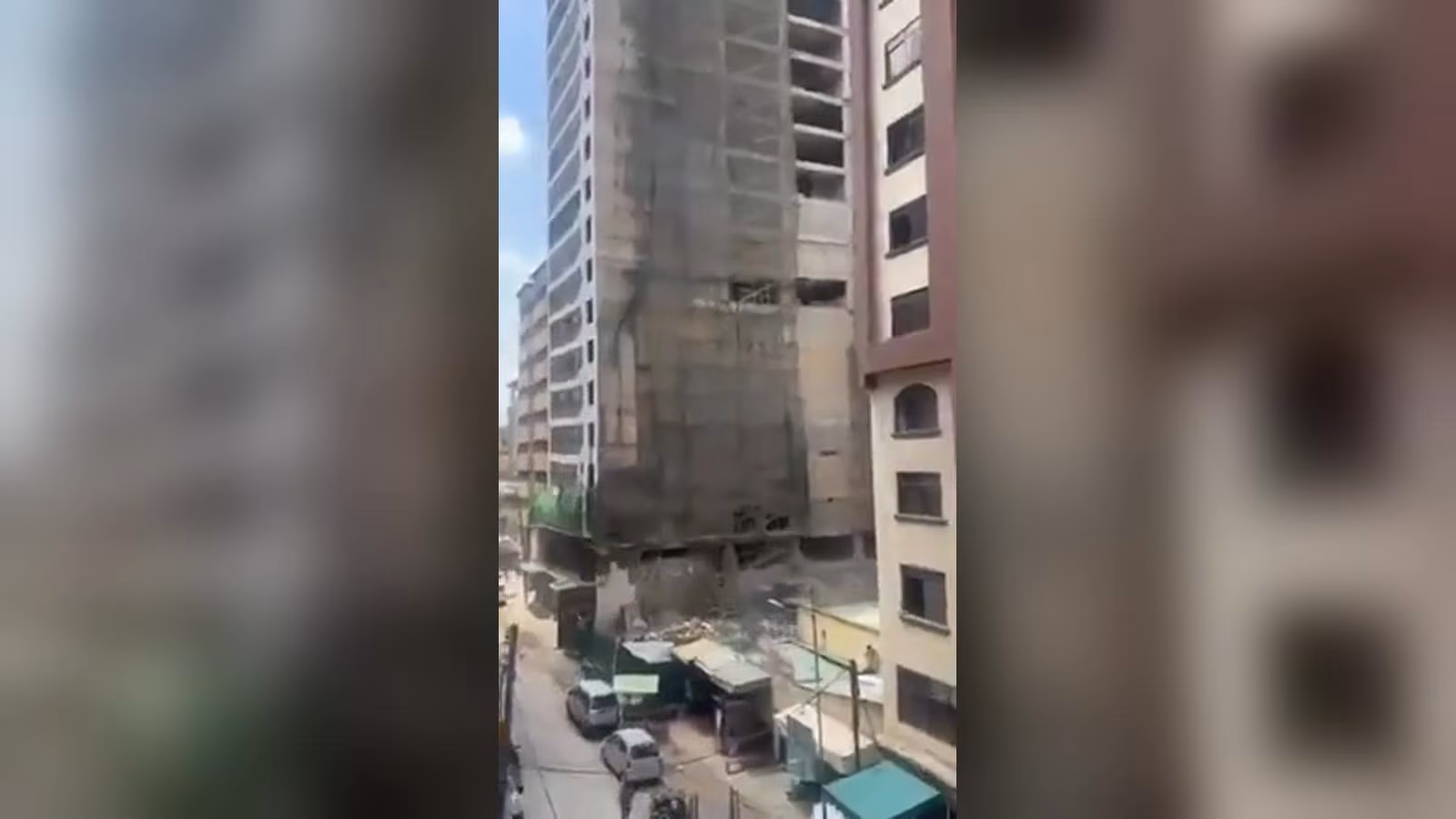 İnşaat iskelesi ve file ile sarılı yarım kalmış yüksek bina; tabanında moloz, sokakta park halinde arabalar.