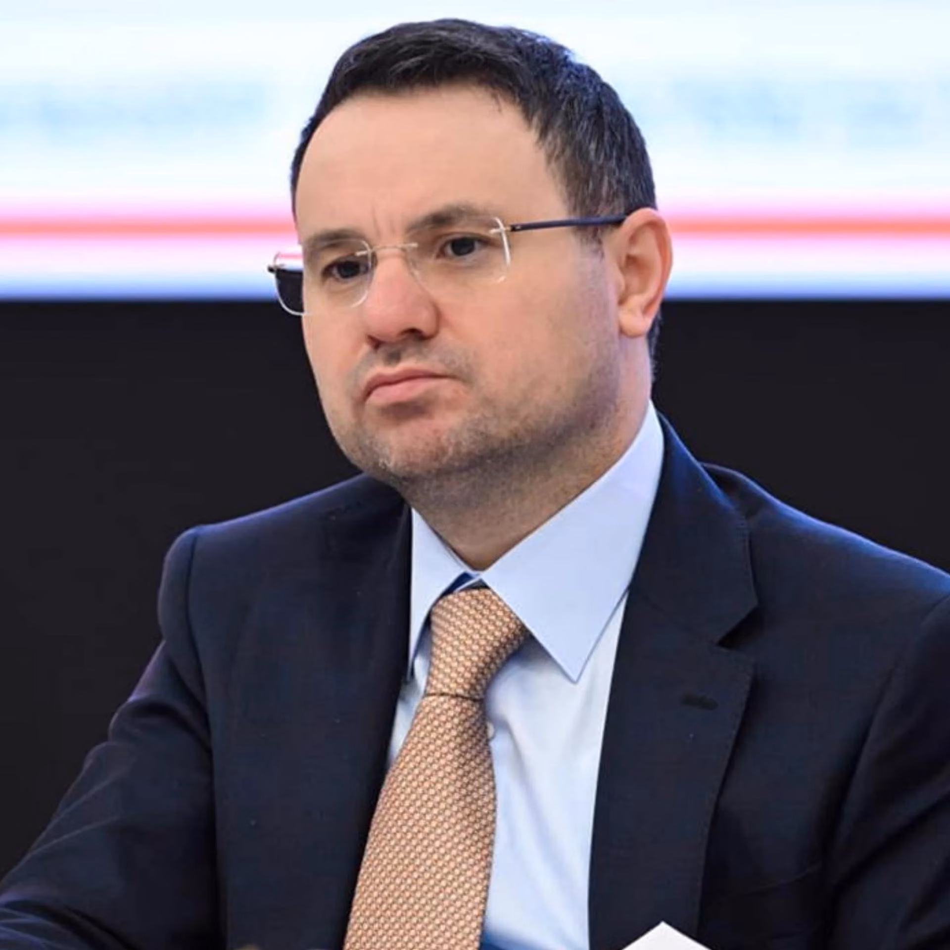 Kısa saçlı, çerçevesiz gözlüklü, lacivert takımlı, açık mavi gömlekli ve bej desenli kravatlı bir erkek.