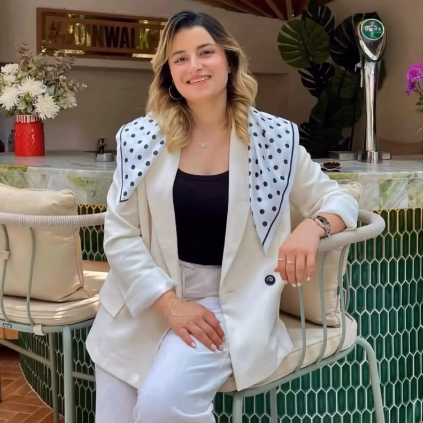 Dalgalı saçlı, beyaz blazer ve puantiyeli eşarp takan bir kadın, yeşil fayanslı barın önündeki koltukta gülümsüyor.